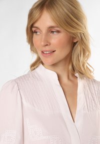 Helles rosa Blusenshirt mit vertikalen Nadelstreifen auf den Schultern, bestickten geometrischen Mustern und einer dezenten strukturierten Oberfläche.