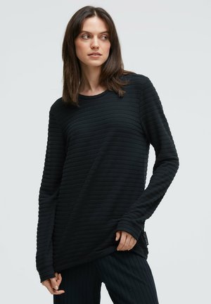 Schwarzes Langarmshirt mit vertikalem Rippenmuster, rundem Ausschnitt und lockerem Sitz. Kombiniert mit dunkel gerippten Hosen, vor neutralem Hintergrund präsentiert.