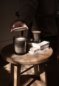 Bougie noire dans un récipient en verre avec couvercle en bois, étiquetée "SKANDINAVISK RO", posée sur un tabouret rond en bois à côté d'une bougie plus petite et d'une pile de livres.