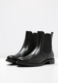 Bottes Chelsea en cuir noir avec panneaux latéraux élastiques, bout rond, texture lisse et petit talon bloc. Design minimaliste sans éléments métalliques visibles.