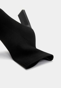 Chaussure à talon haut de style chaussette noire avec une tige en tissu texturé et un talon carré. Le design est élégant avec une silhouette épurée.