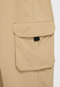 Spodnie cargo w kolorze khaki o fakturze materiału, wyposażone w dużą klapkową kieszeń i czarny pasek wzmacniający. Prosty, funkcjonalny design.