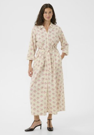 Žena v dlhých béžových šatách s ružovými kvetinovými vzormi, trištvrťovými rukávmi, golierom a pásikom v páse, v kombinácii s čiernymi T-pásikovými lodičkami.