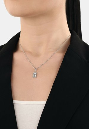Collana in argento con un pendente rettangolare caratterizzato da un design intricato, indossata sopra una maglietta bianca e sotto un blazer nero.