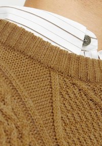 Suéter marrón de punto con patrón texturizado y cuello acanalado, superpuesto a una camisa blanca con cuello y finas franjas marrones y botón.