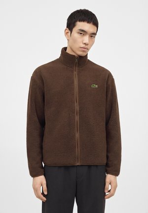 Homme portant une veste polaire marron zippée avec un petit logo crocodile vert sur la poitrine, debout devant un fond uni.