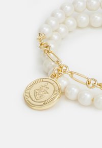 Bracciale di perle con dettagli in catena tono oro e un ciondolo circolare caratterizzato da un'emblema in rilievo. Perle lisce e lucide di colore bianco.