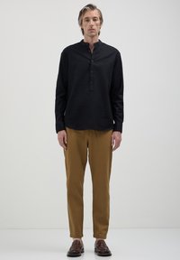 Homme se tenant contre un fond uni, portant une chemise noire à boutons, un pantalon marron et des chaussures en cuir marron, faisant face avec les mains détendues.