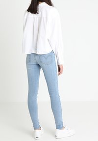 Mujer vistiendo jeans ajustados de color azul claro, camisa blanca de botones y zapatillas blancas, de pie contra un fondo blanco liso, vista desde atrás.
