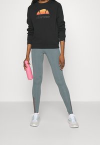 Personne portant un sweat-shirt noir avec un logo orange, des leggings gris avec des panneaux en mesh, des baskets blanches, tenant une bouteille d'eau rose à la main.