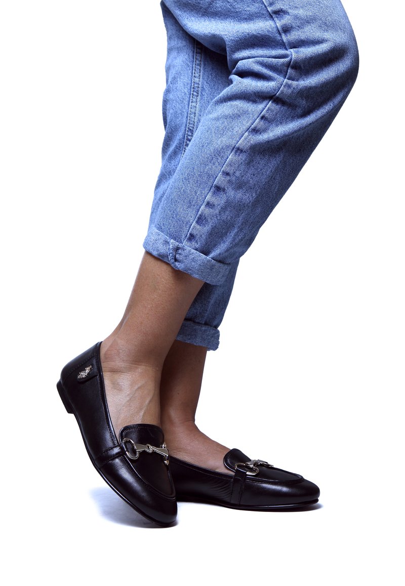 Mocassini in pelle nera con un accento di catena argentata; suola piatta, punta rotonda, abbinati a jeans in denim azzurro chiaro arrotolati.