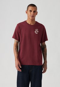 Burgunderfarvet bomulds T-shirt med rund hals, korte ærmer og et trægrafik på brystet. Båret med mørke denimjeans.