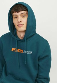 Sudadera color azul verdoso hecha de un tejido suave, con un bolsillo tipo canguro en el frente y un logotipo en naranja y blanco de "JACK & JONES" en el pecho.