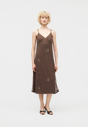 Γυναίκα που φοράει καφέ midi φόρεμα χωρίς μανίκια με λεπτές τιράντες και διακριτικά κεντημένα λουλουδένια μοτίβα, σε συνδυασμό με μαύρα πέδιλα ανοιχτής μύτης.