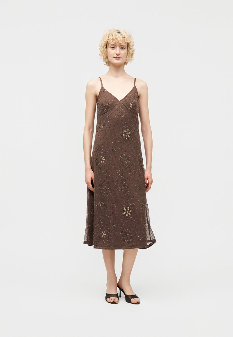 Γυναίκα που φοράει καφέ midi φόρεμα χωρίς μανίκια με λεπτές τιράντες και διακριτικά κεντημένα λουλουδένια μοτίβα, σε συνδυασμό με μαύρα πέδιλα ανοιχτής μύτης.