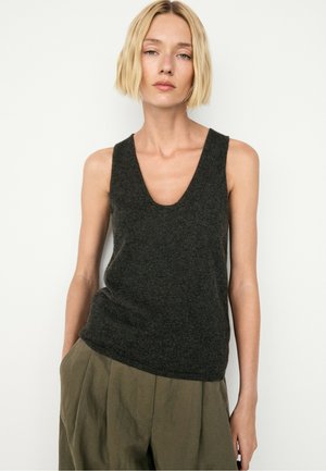 Donna bionda che indossa un top senza maniche grigio scuro con scollo a V e pantaloni larghi verde oliva, in piedi di fronte a uno sfondo bianco.