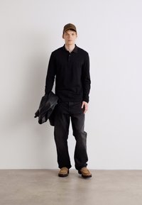 Μαύρο πουκάμισο μακρυμάνικο πόλο, σκούρα τζιν, καφέ παπούτσια και σκούρο μπουφάν που κρατιέται στο αριστερό χέρι. Καφέ καπέλο με λογότυπο. Ουδέτερο φόντο.