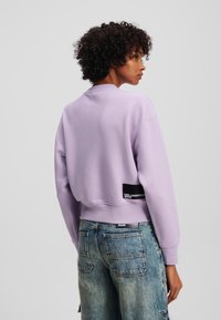 Lavender cropped sweatshirt with a round neck and ribbed cuffs, featuring a black logo patch at the back and paired with faded denim.