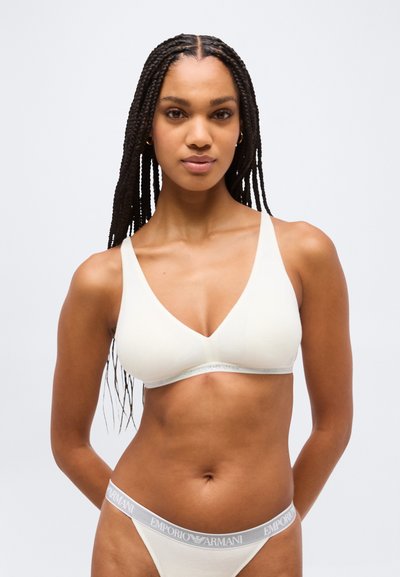 Femme aux longues tresses portant un soutien-gorge blanc à encolure en V et une culotte assortie avec une ceinture Emporio Armani, debout devant un fond uni.