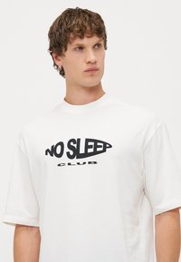 Biele tričko s krátkym rukávom, na ktorom je v tučných čiernych písmenách nápis „NO SLEEP CLUB“. Je vyrobené z mäkkého materiálu, má voľný strih a okrúhly výstrih.