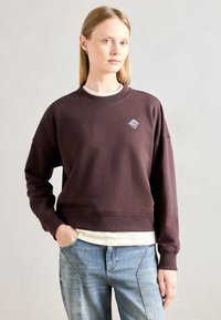 Sudadera marrón con cuello redondo y puños de canalé. Cuenta con un pequeño logo blanco en el pecho. Llevada sobre una camiseta de color crema.