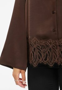 Brun bluse med lange ermer, med en teksturert blonderkant nederst. Stoffet er glatt med en subtil glans.