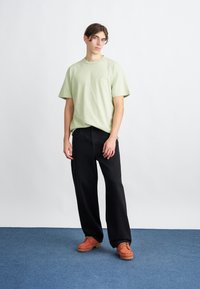 Camiseta verde claro de manga corta, pantalones negros de pierna ancha y zapatos naranjas con cordones. Diseño minimalista, texturas lisas y detalle de marca sutil.