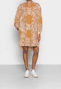 Langærmet kjole i sennepsfarve med et hvidt blomstret paisley-mønster, med V-hals og afslappet pasform, båret med hvide sneakers.