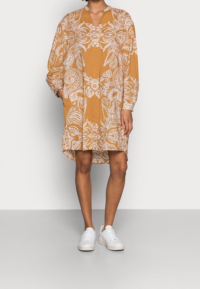 Langærmet kjole i sennepsfarve med et hvidt blomstret paisley-mønster, med V-hals og afslappet pasform, båret med hvide sneakers.