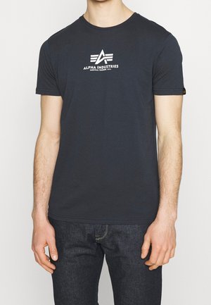 Homme portant un t-shirt noir à manches courtes avec le logo blanc Alpha Industries et un texte, associé à un jean bleu foncé.