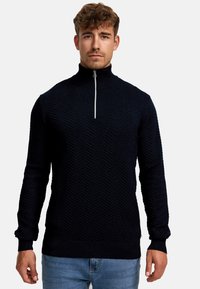 Dunkelblauer gestrickter Pullover mit hohem Kragen, der ein strukturiertes Zickzackmuster und einen silbernen Reißverschluss am Hals aufweist.