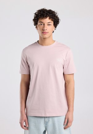 Jonge man met krullend haar, die een lichtroze T-shirt en lichtblauwe spijkerbroek draagt, staand tegen een effen lichtgrijze achtergrond.