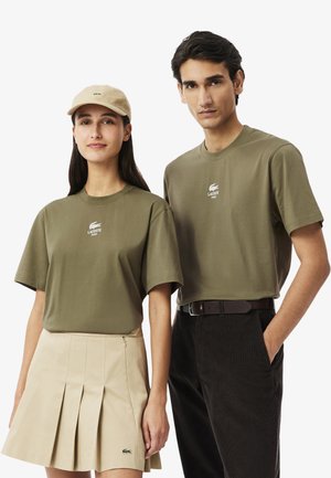 Olijfgroene katoenen T-shirts met korte mouwen en logo, gecombineerd met een beige plissérok en donkere flanelle pantalon. Beige pet inbegrepen.