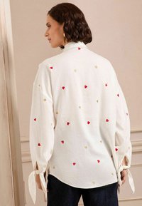 Blusa bianca a maniche lunghe con motivi di cuori rossi e dorati, dotata di lacci ai polsini e dalla vestibilità ampia, vista da dietro.