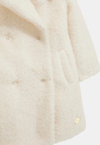 Cappotto morbido color crema con design doppiopetto, quattro bottoni decorativi e un accento logo in tono oro sulla parte anteriore inferiore.