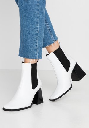 Hvide ankelstøvler med sorte elastiske sidelommer og chunky hæle båret med cropped blå cowboybukser på gråt gulv.