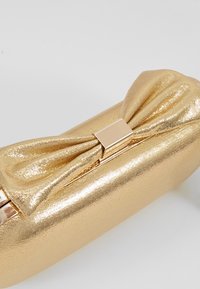 Zlatá metalická kabelka ve tvaru clutche s detailem ve tvaru mašle a obdélníkovým zapínáním na bílém pozadí.