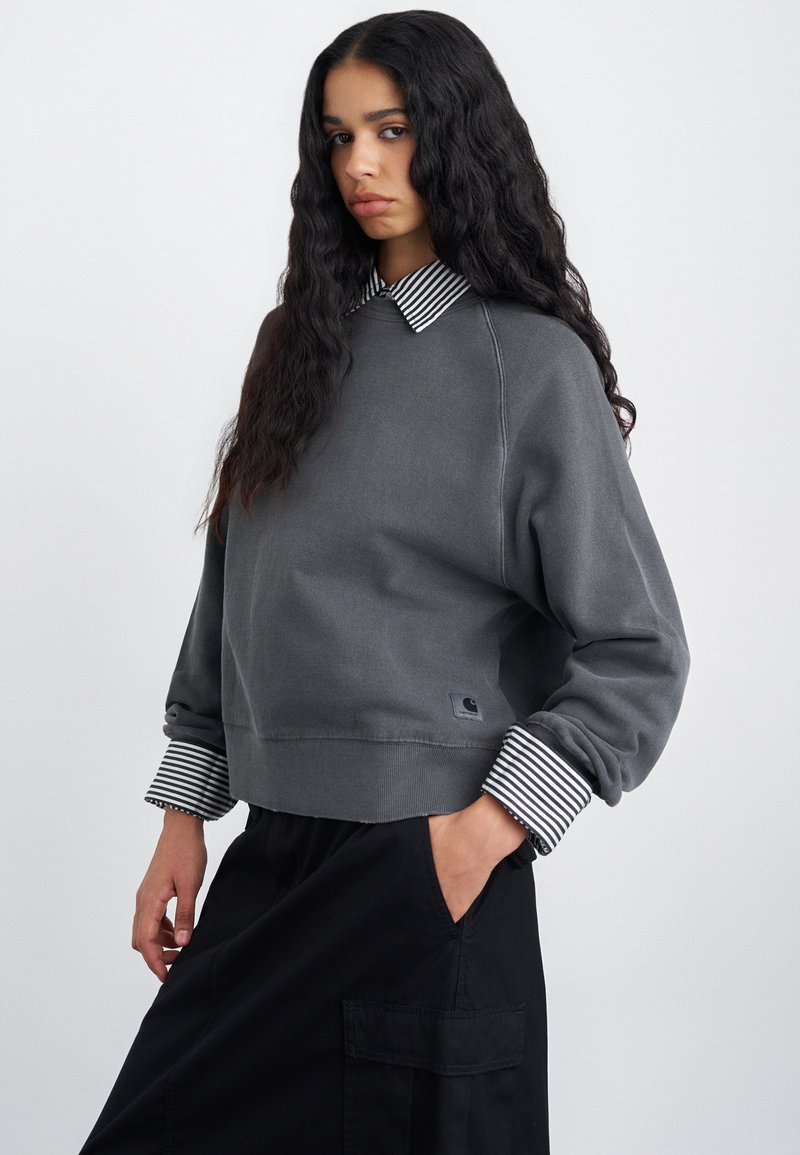 Sivý sveter s pruhovanými manžetami a golierom, s voľným strihom a malým logom značky v dolnom ľavom rohu. Čierne nohavice sú čiastočne viditeľné.