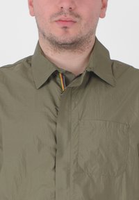 Camicia leggera color verde oliva con una texture stropicciata e colletto a bottoni. Presenta una tasca sul petto sinistro e un dettaglio della cerniera multicolore.