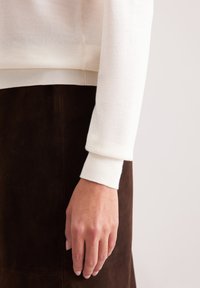 Ivory wełniany sweter o miękkiej fakturze, z prążkowanymi ściągaczami. W połączeniu z brązową zamszową spódnicą, ukazującym zbliżenie na ramię i dłoń.