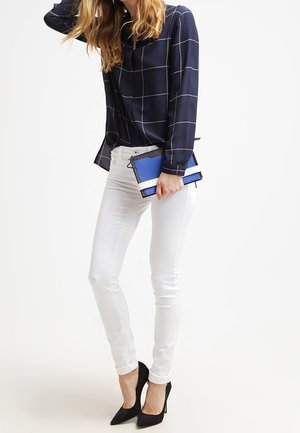 Femme portant une blouse à carreaux bleu marine et un pantalon blanc, tenant une pochette bleue et blanche, portant des escarpins noirs, se tenant devant un fond blanc.