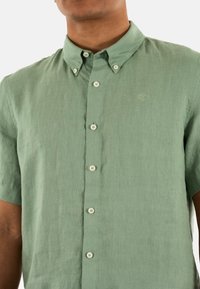 Camicia a manica corta, con bottoni, in lino verde chiaro. Presenta un colletto, cinque bottoni e un logo sottile sul petto.