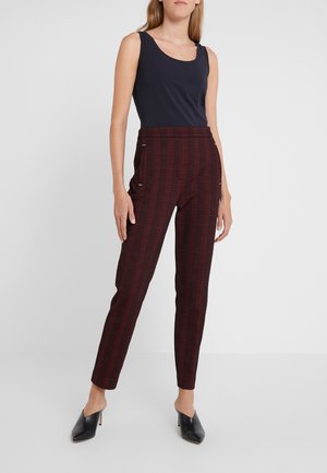 Kobieta w czarnej bluzce bez rękawów, bordowych spodni w kratę z wysokim stanem oraz czarnych, szpiczastych mule na obcasie, stojąca na tle gładkiej białej ściany.