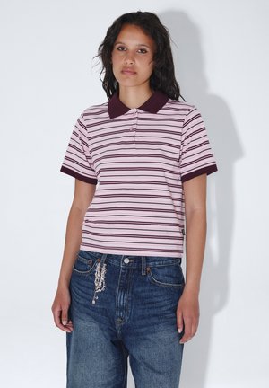Jong persoon met middellang donker haar die een roze gestreept poloshirt en blauwe spijkerbroek met een kralenketting als accessoire draagt.