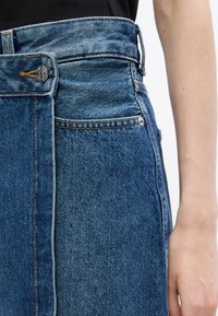 Denimrock mit hoher Taille, Knopfverschluss, einer vorderen Tasche und sichtbaren Nähten in einem mittleren Blauton.