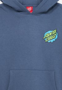 Kék kapucnis pulóver kenguru zsebbel és bordázott mandzsettákkal. A bal mellrészen színes "Santa Cruz Skateboards" grafika található.