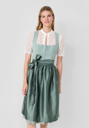 Frau trägt ein hellgrünes traditionelles Kleid mit einer strukturierten weißen Bluse und einem gebundenen Satin-Schürzenrock, stehend vor einem schlichten Hintergrund.
