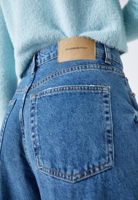 Los pantalones vaqueros de mezclilla azul cuentan con un diseño estándar de cinco bolsillos, con costuras naranjas y un parche de logo de cuero marrón en la cintura trasera.