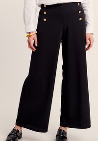 Zwarte wijde broek van zachte stof, met gouden knopen op de tailleband en een gladde textuur. Gemaakt in combinatie met een wit shirt.