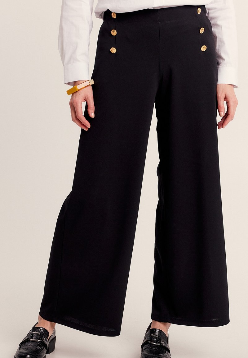 Zwarte wijde broek van zachte stof, met gouden knopen op de tailleband en een gladde textuur. Gemaakt in combinatie met een wit shirt.
